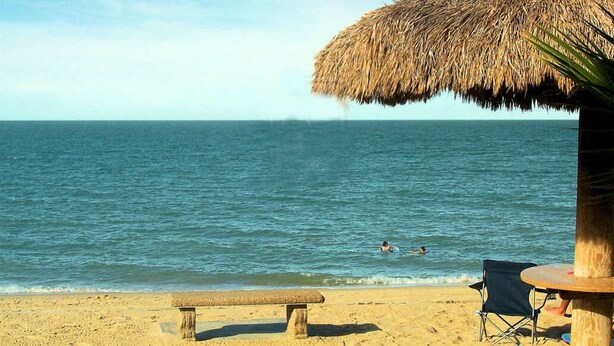 Ein Strand mit einem Strohdachschirm, einer Sitzbank und einem Tisch mit einem Stuhl.
