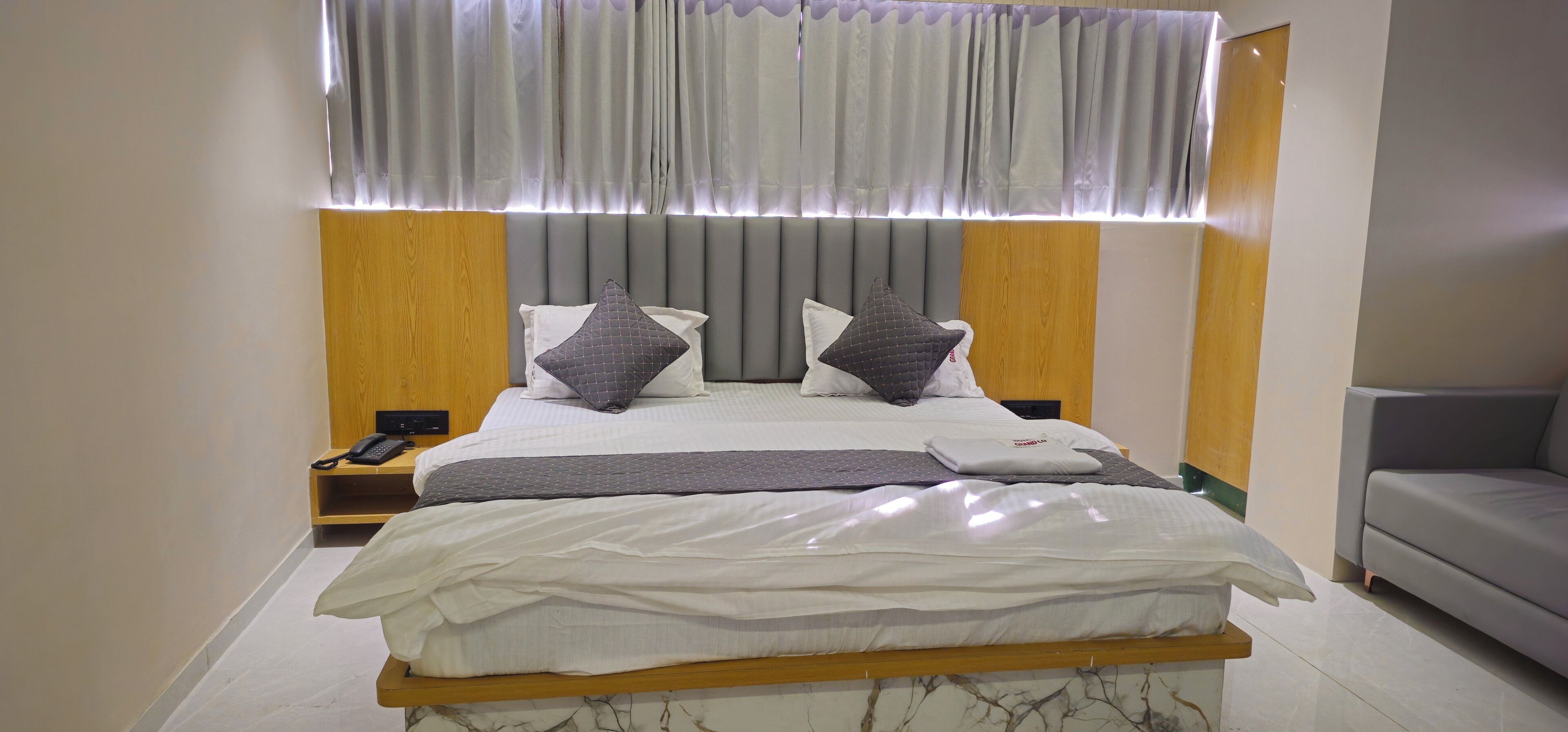 A modern hotel room with a large bed, a bedside table with a phone, and a grey sofa.