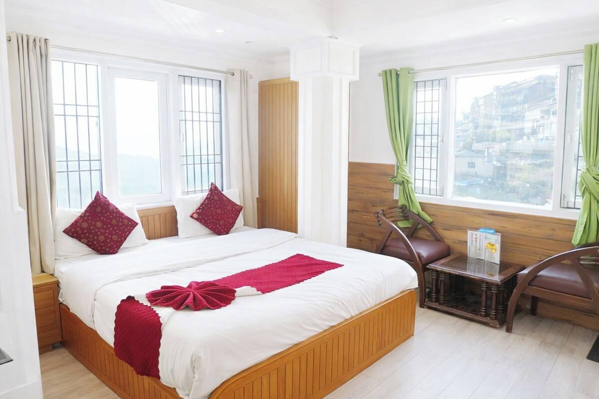 Ein Hotelzimmer mit einem großen Bett, einem Holzkopfende, einem kleinen Tisch und Blick auf die Stadt durch die Fenster.