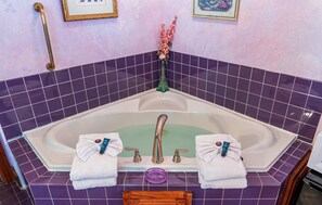 Une salle de bain avec une grande baignoire, des carreaux violets, deux serviettes, un vase vert et un tableau encadré au mur.