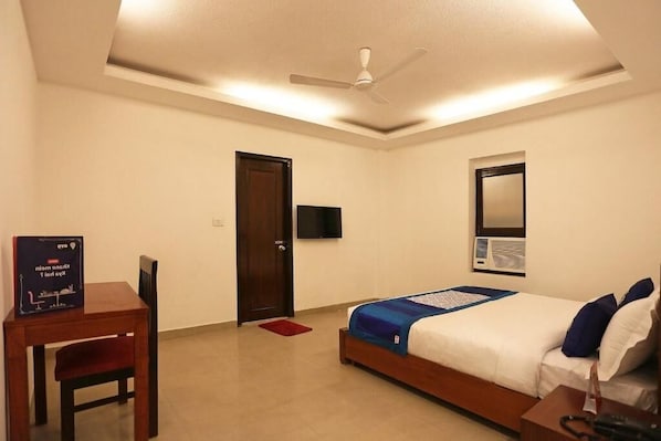 Een hotelkamer met een bed, een bureau, een stoel, een plafondventilator, een televisie en een raam.