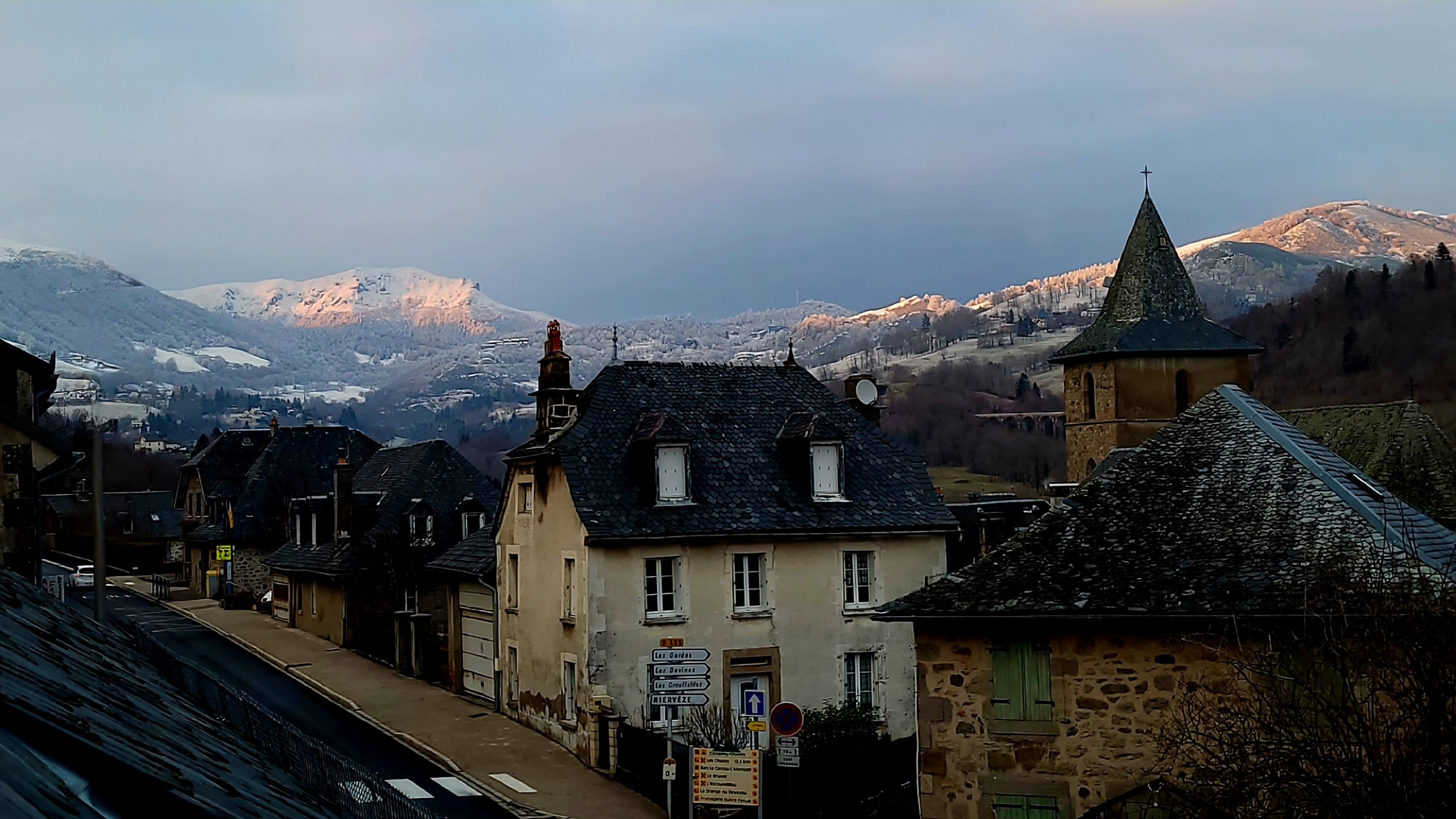 Eine Stadt mit historischen Steinbauten, einer Kirche mit spitzem Kirchturm und verschneiten Bergen im Hintergrund.