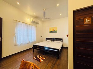 A hotel room with a bed, a ceiling fan, an air conditioning unit, a window with curtains, and a door with a sign.