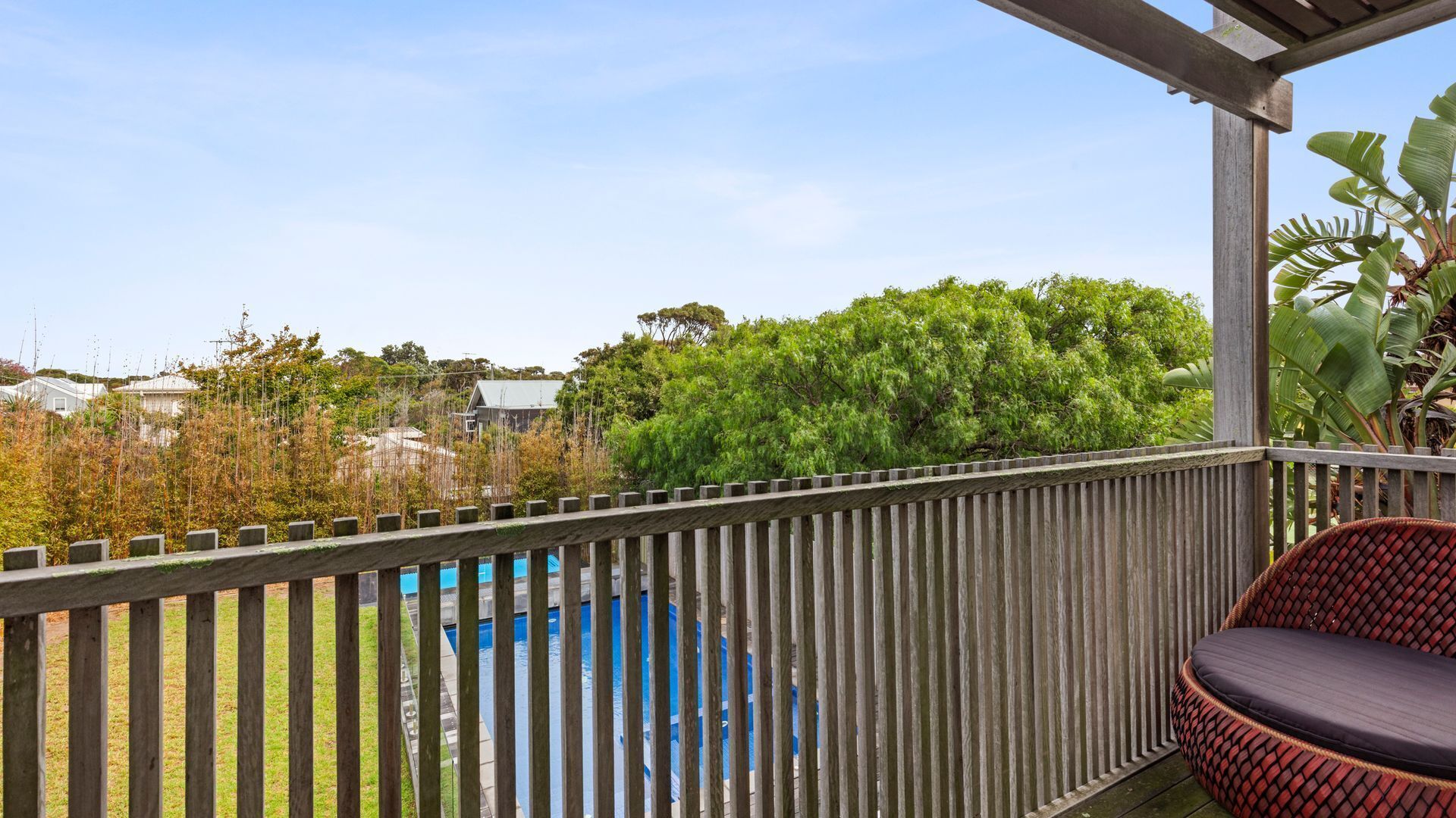 A wooden deck with a pool, a wicker chair, and a view of greenery and houses.