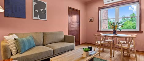 A living room with a sofa, a coffee table, a dining table with chairs, a window with a view, and two wall art pieces.