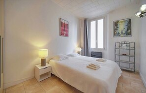 Ein Hotelzimmer mit einem Bett, zwei Nachttischlampen, einem Fenster mit Vorhängen, einem Metallregal und zwei Bildern an der Wand.