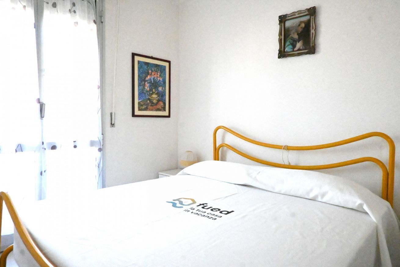 A bed with a logo, a framed painting, and a small table with a lamp.