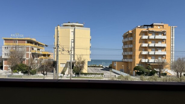 A multi-story building with balconies, a clear sky, and a distant view of the sea.