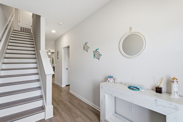 Uma escadaria leva a um corredor com um console branco, um espelho redondo e obras de arte decorativas na parede.