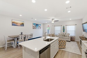 A modern kitchen with a central island, white cabinetry, and a dining area with a table and chairs. - Ocean Breeze Stay and Play! 3BR + Game Room (Panama City Beach)