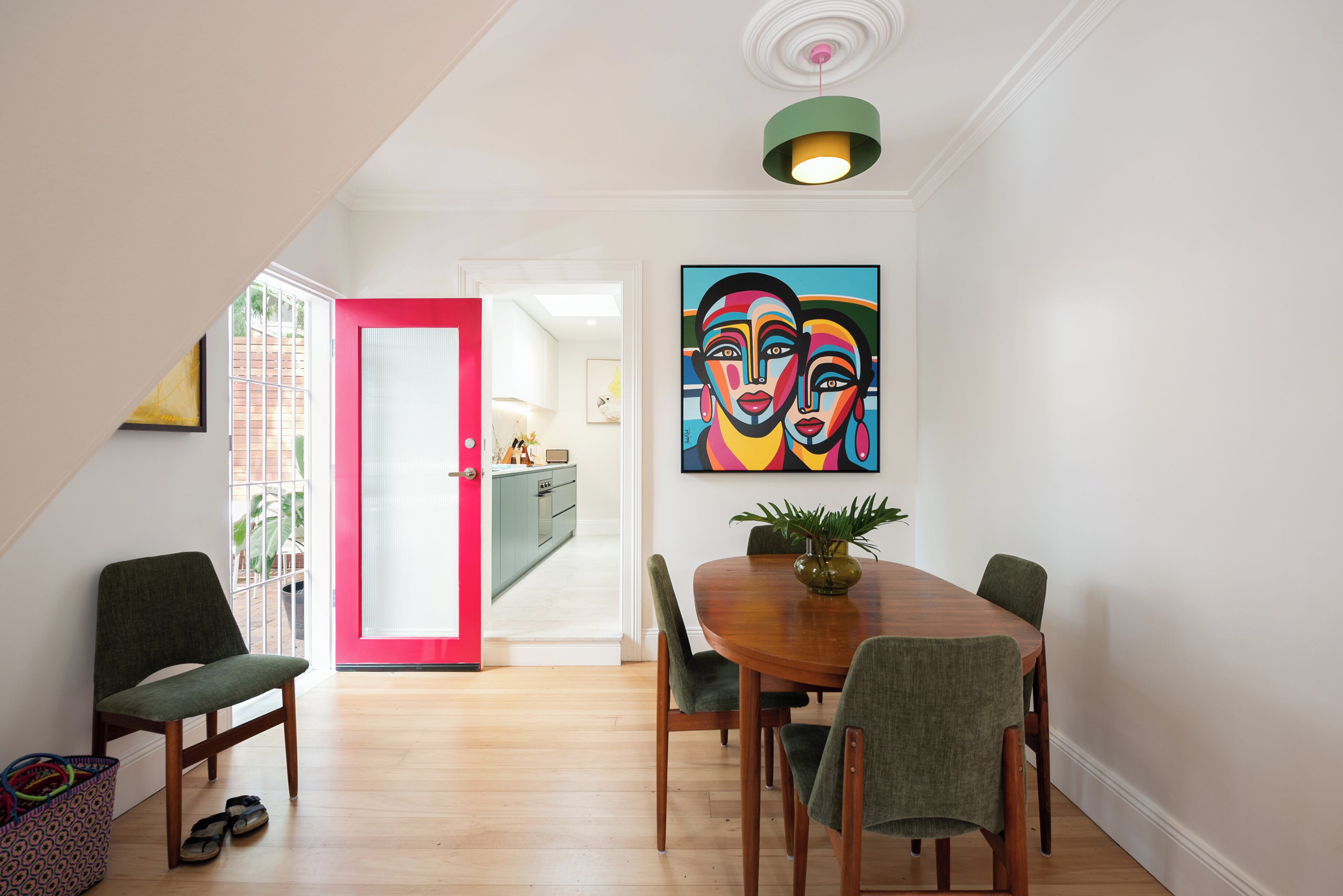 A dining area with a wooden table, green upholstered chairs, a colorful abstract painting, and a bright red door.