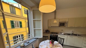 Ein Essbereich mit einem Tisch für vier Personen, ein Fenster mit Blick auf ein gelbes Gebäude und eine Küche mit weißen Schränken und einer Mikrowelle.