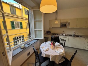 Ein Essbereich mit einem Tisch für vier Personen, ein Fenster mit Blick auf ein gelbes Gebäude und eine Küche mit weißen Schränken und einer Mikrowelle.