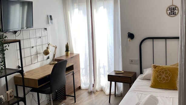 Un dormitorio con cama, escritorio, silla, televisión y ventana con cortinas.