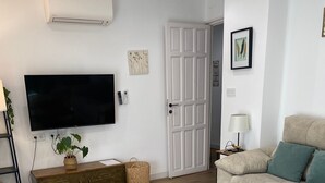Una sala de estar con una televisión de pantalla plana, un sofá, una mesa de centro, una silla, una lámpara y una puerta.