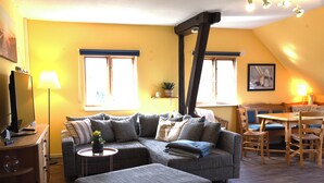 A living room with a sectional sofa, a coffee table, a TV, a lamp, and a dining area with a table and chairs. - Cozy vacation apartment on the Baltic Sea, with large garden (Behrensdorf (Ostsee))