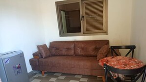 Un canapé marron, une petite table recouverte d’une nappe à motifs, un réfrigérateur et une fenêtre avec des stores.