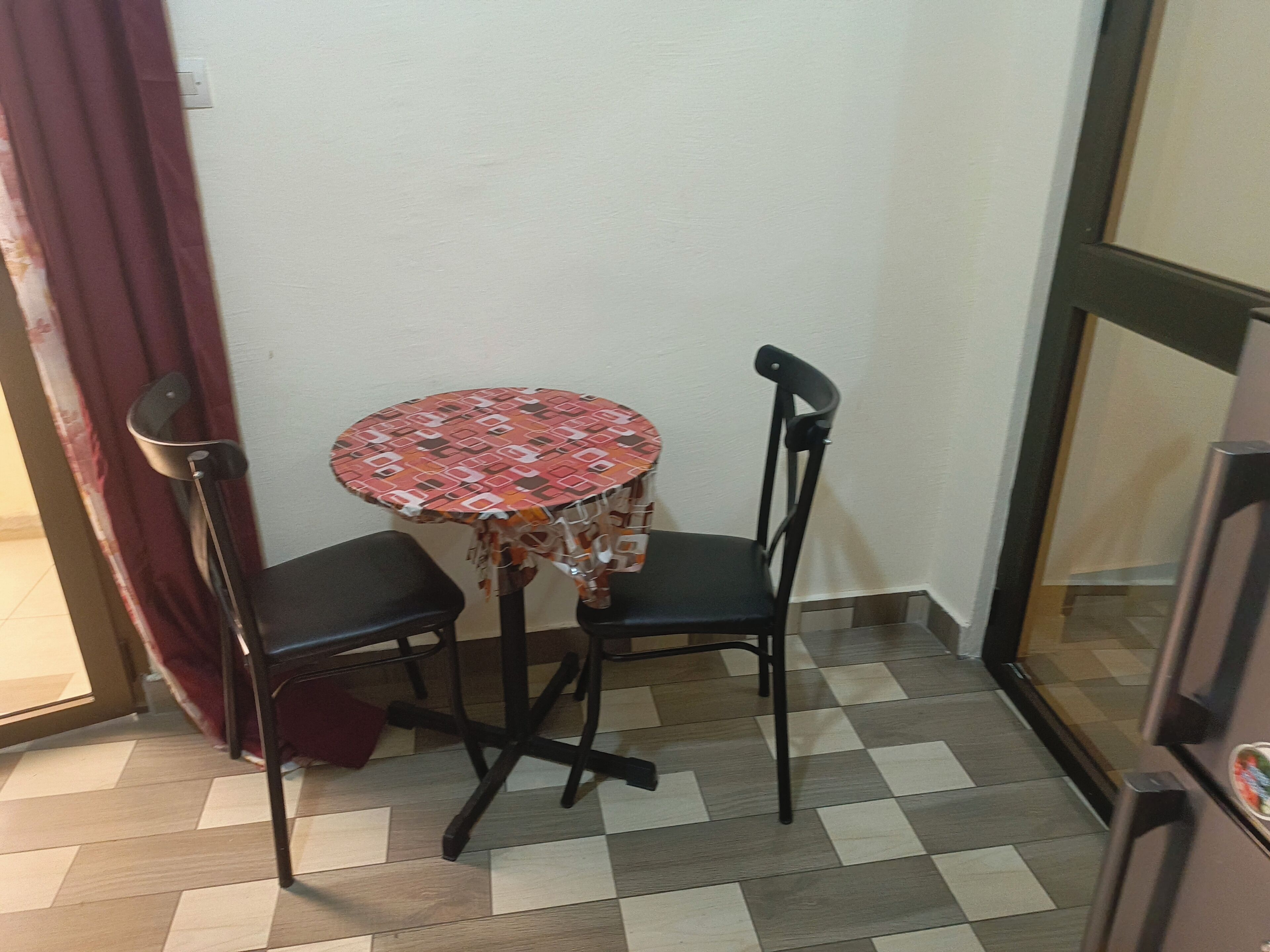 Una habitación pequeña con una mesa redonda, dos sillas, una cortina y un refrigerador.