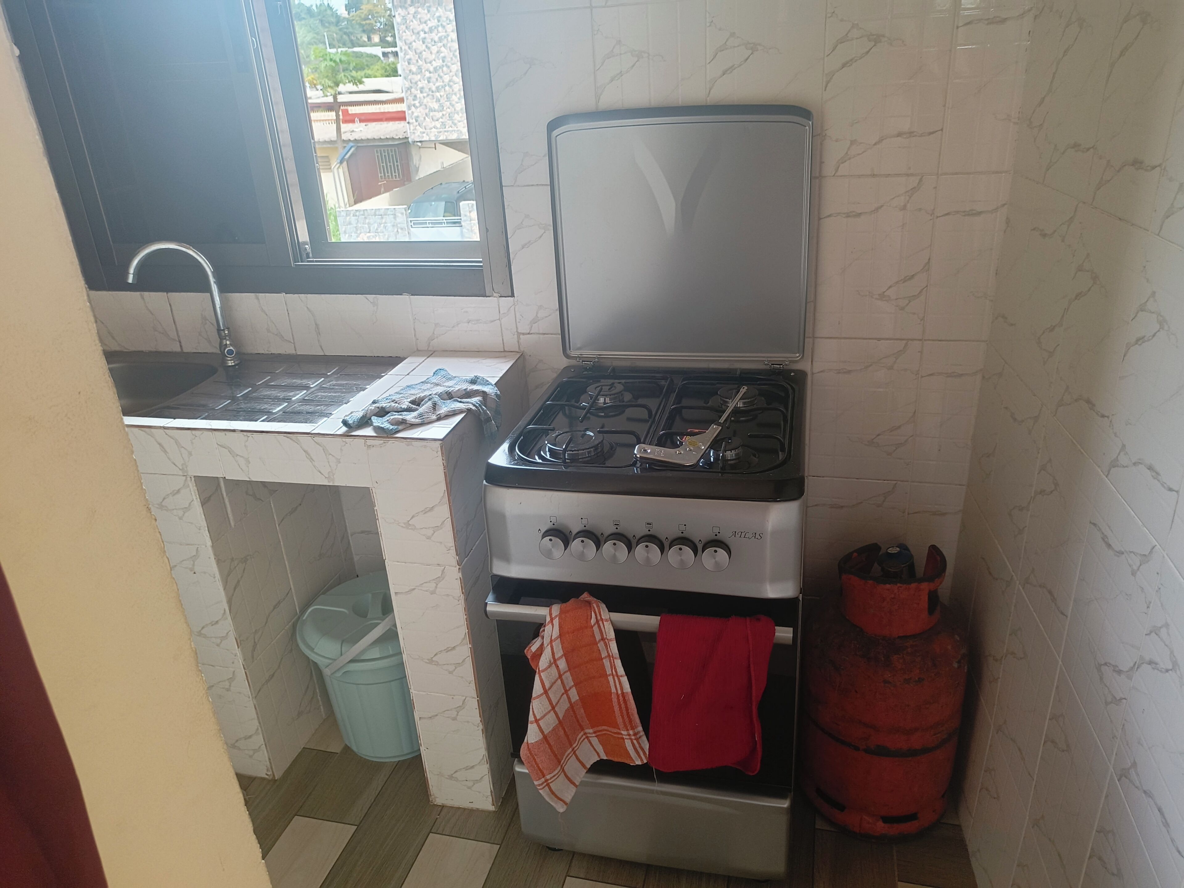 A kitchen with a gas stove, a sink, a window, a trash bin, and a gas cylinder.