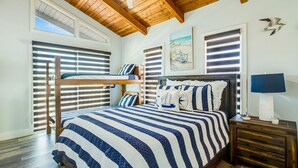 Ein Schlafzimmer mit Etagenbett, einem großen Bett mit gestreifter Bettwäsche, einem Deckenventilator, Holzbalken und einem Strandgemälde.