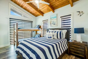 Ein Schlafzimmer mit Etagenbett, einem großen Bett mit gestreifter Bettwäsche, einem Deckenventilator, Holzbalken und einem Strandgemälde.