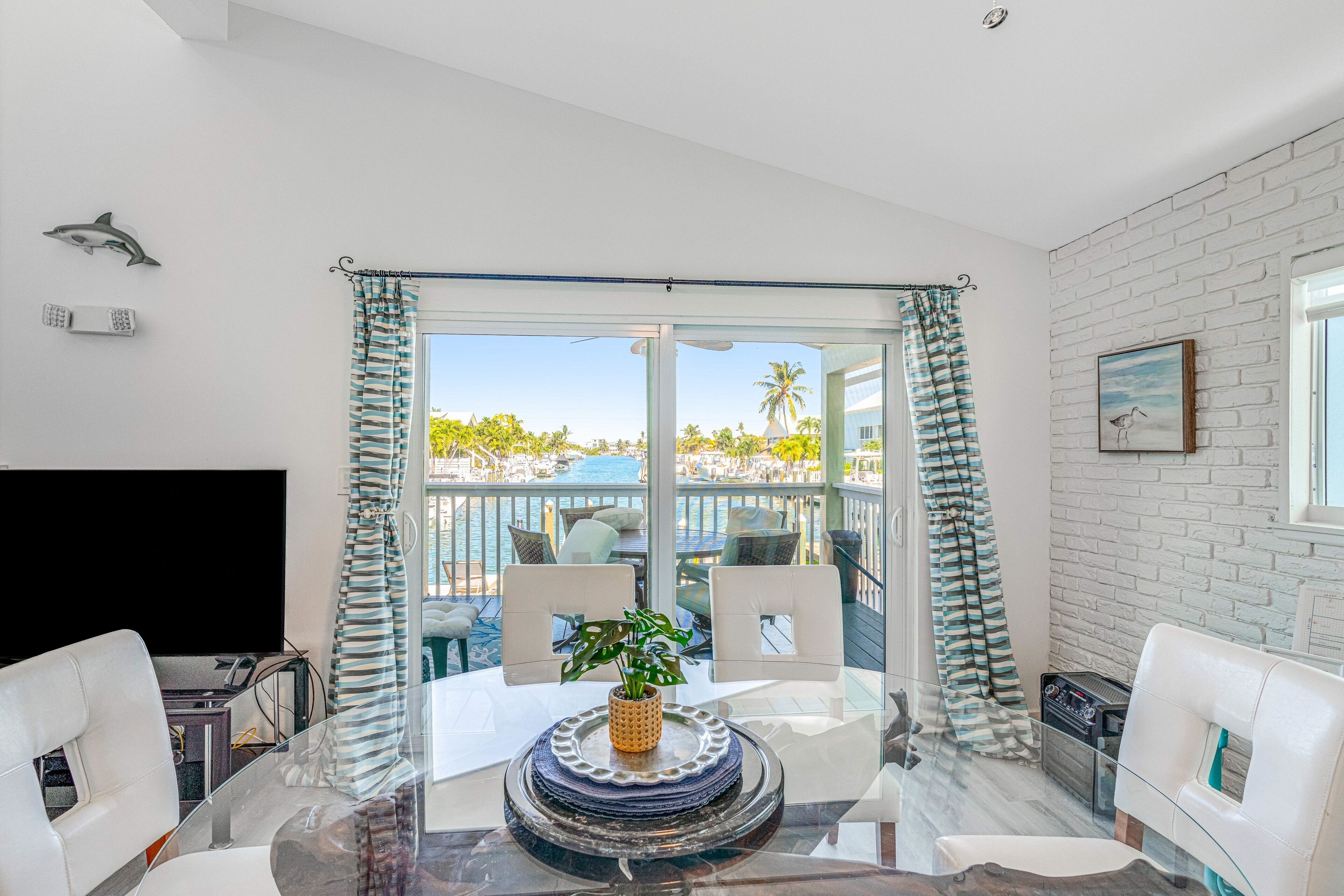 A modern room with a glass table, white chairs, a TV, and a view of a pool and palm trees.