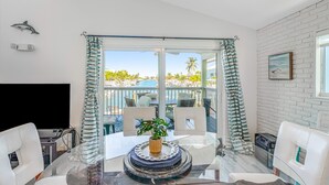 Une chambre moderne avec une table en verre, des chaises blanches, un téléviseur et une vue sur une piscine et des palmiers.