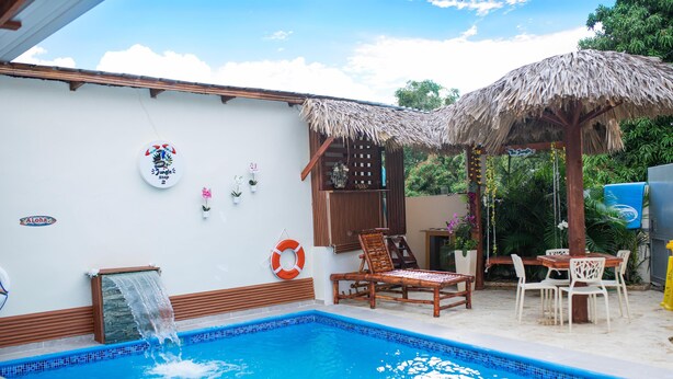 A swimming pool with a waterfall feature, wooden lounge chairs, and a thatched roof structure. - The Jungle: Private Tropical Villa : Pool & 6BR (Puerto plata)