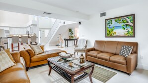 A spacious living room with a brown leather sofa, a glass coffee table, and a large painting of a beach scene. - Waterfront home with amazing views, boat dock, screened porch & water sport gear (Key Largo)