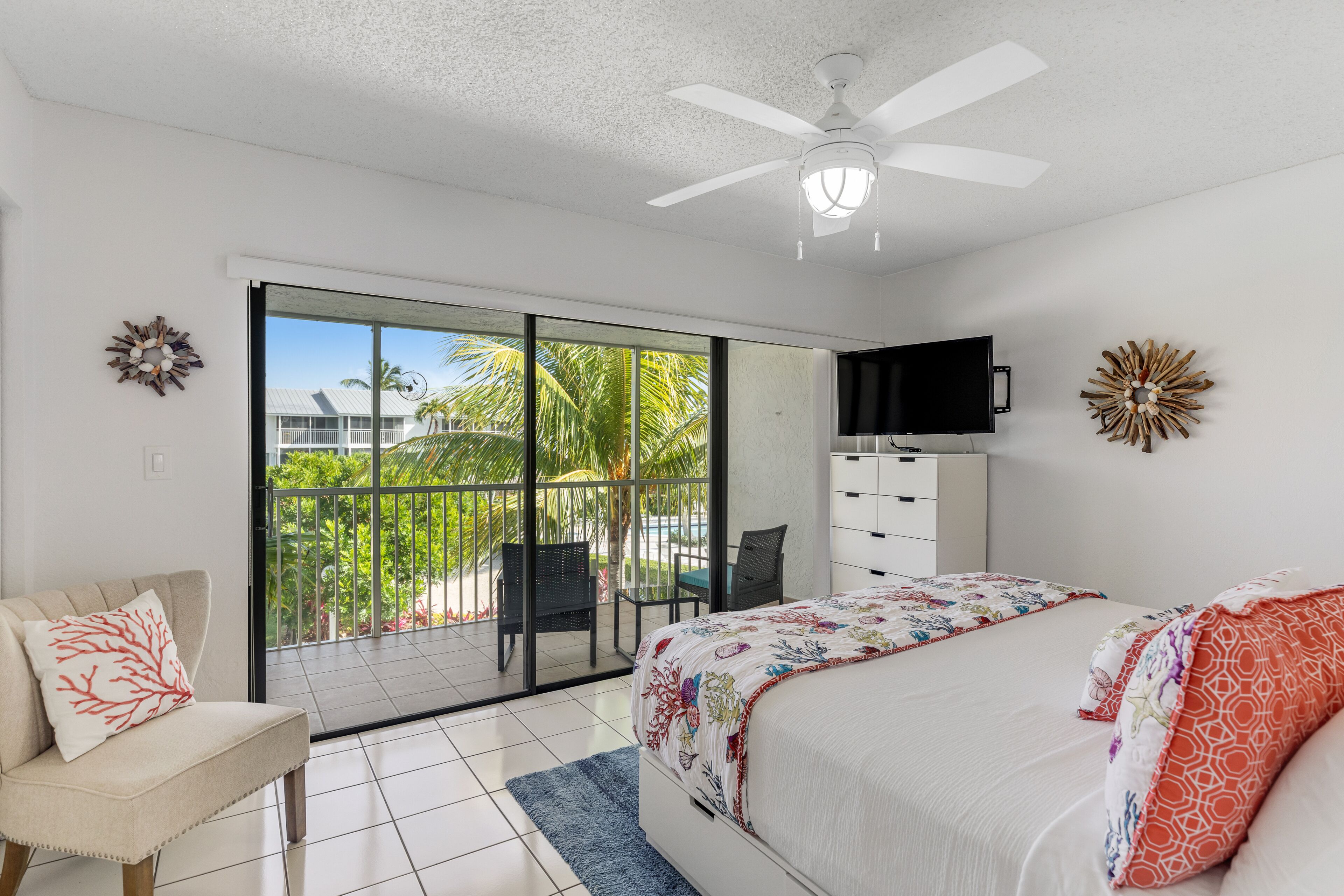 A bedroom with a bed, a TV, a ceiling fan, and a balcony with a chair and a table.