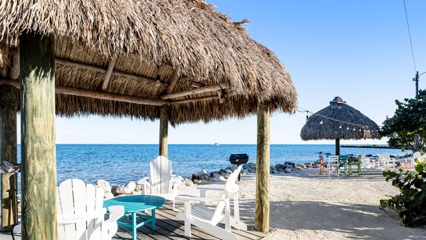 A beachside deck with thatched-roof structures, white chairs, and a view of the sea. - Tropical condo with 2 pools, private beach area, swimming lagoon, tennis & AC (Key Largo)
