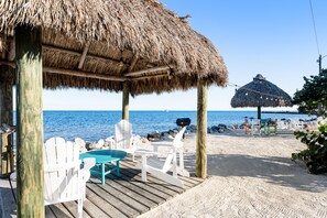 A beachside deck with thatched-roof structures, white chairs, and a view of the sea. - Tropical condo with 2 pools, private beach area, swimming lagoon, tennis & AC (Key Largo)