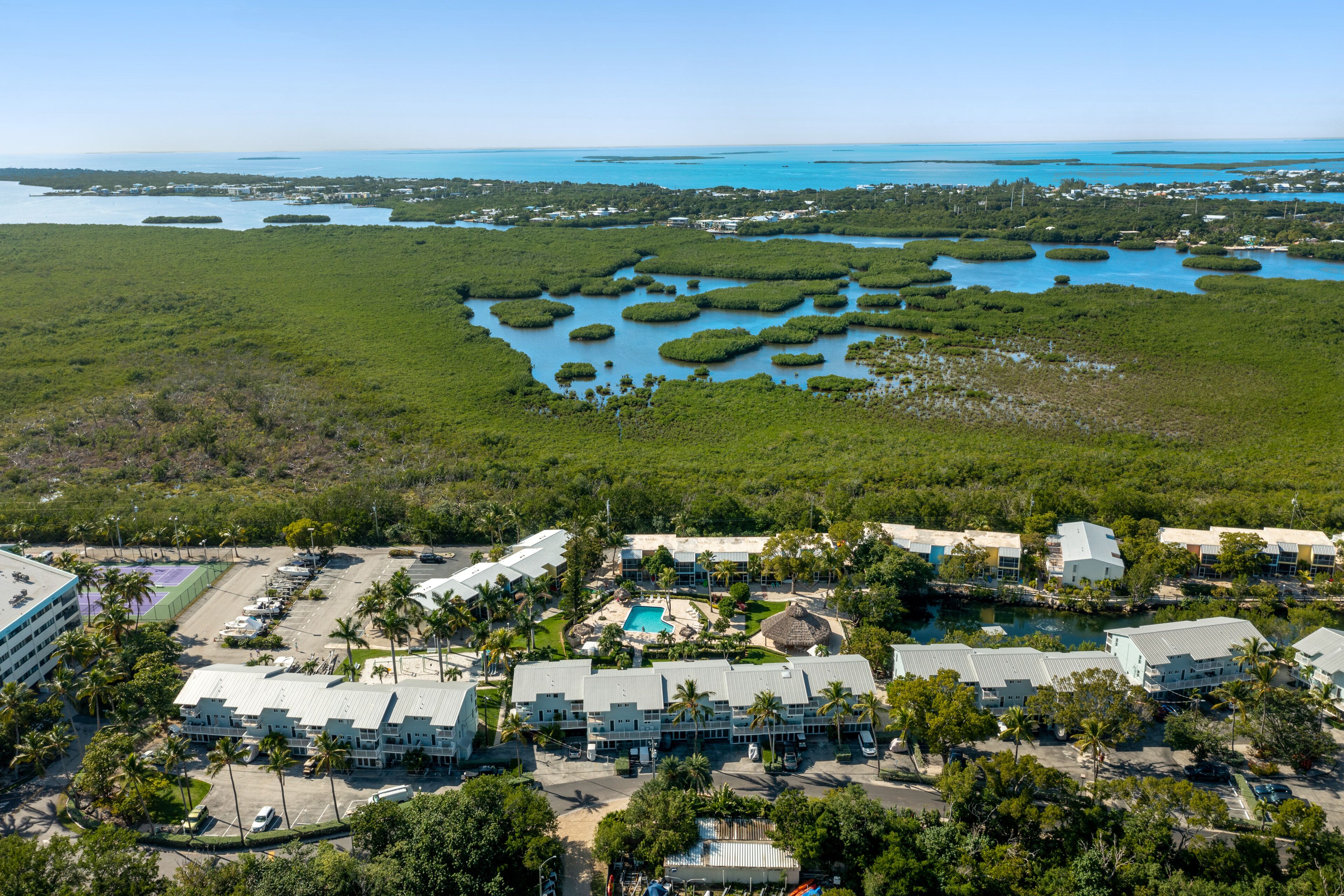 Vue aérienne d’un complexe hôtelier comprenant plusieurs bâtiments, une piscine et une étendue d’eau bordée de mangroves.