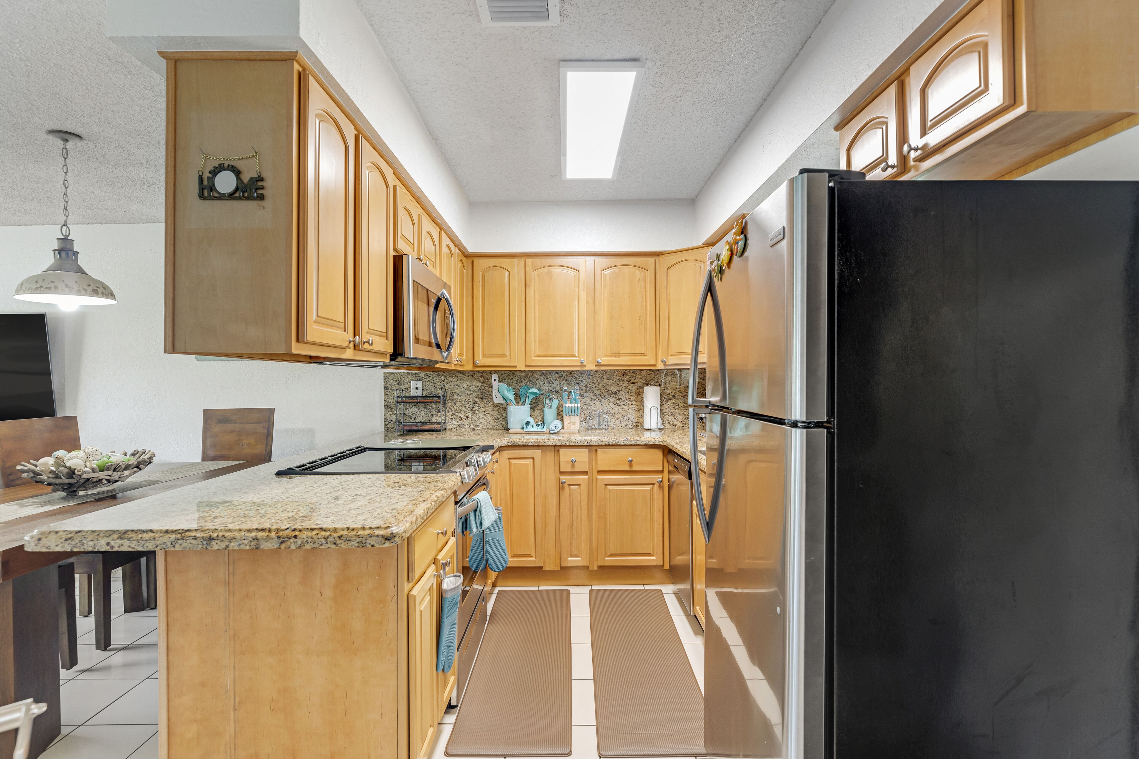 A kitchen with wooden cabinets, a black refrigerator, granite countertops, a microwave, and a hanging light fixture.