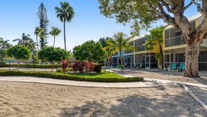A hotel with a modern design, featuring a large tree in the foreground, a well-maintained garden with various plants, and a clear blue sky. - Tropical condo with 2 pools, private beach area, swimming lagoon, tennis & AC (Key Largo)