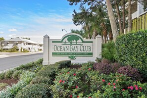Schild für den Ocean Bay Club in der South Ocean Boulevard 201, umgeben von üppiger Vegetation und blauem Himmel.