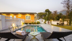 A swimming pool with lounge chairs, a small table, and a wooden building with a white fence. - Alaya Stays Baramasa 3 BHK Hill Retreat (Dehradun)