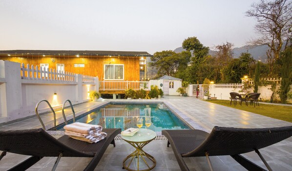 A swimming pool with lounge chairs, a small table, and a wooden building with a white fence. - Alaya Stays Baramasa 3 BHK Hill Retreat (Dehradun)