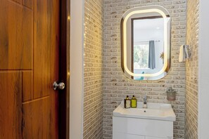 A bathroom with a wooden door, a white sink with two bottles, a mirror with a light, and a brick-patterned wall. - Alaya Stays Baramasa 3 BHK Hill Retreat (Dehradun)