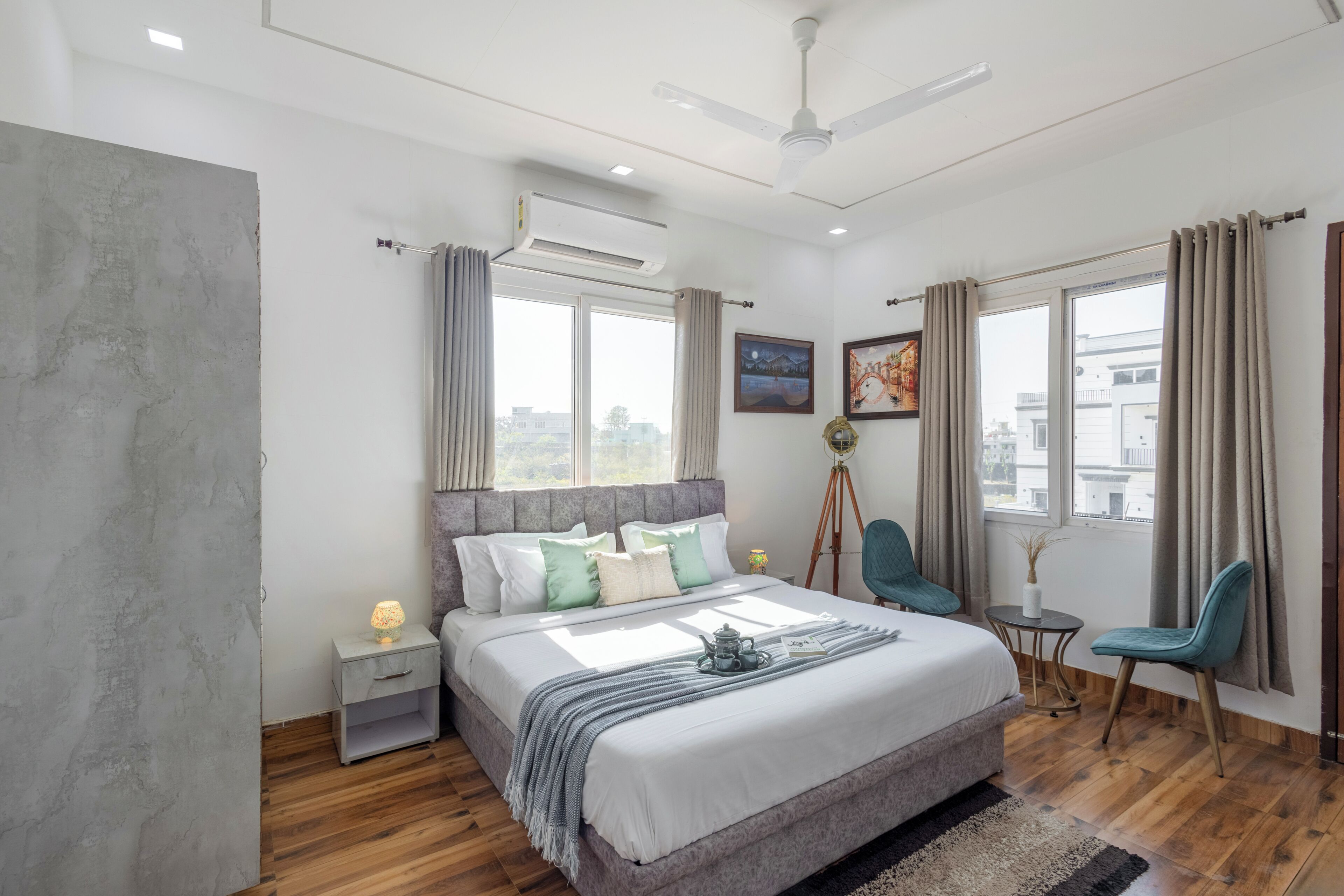 A modern bedroom with a large bed, a ceiling fan, an air conditioning unit, a tripod lamp, and a view of the city through the window.