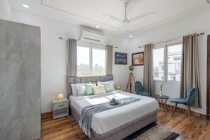 A modern bedroom with a large bed, a ceiling fan, an air conditioning unit, a tripod lamp, and a view of the city through the window. - Alaya Stays Baramasa 3 BHK Hill Retreat (Dehradun)