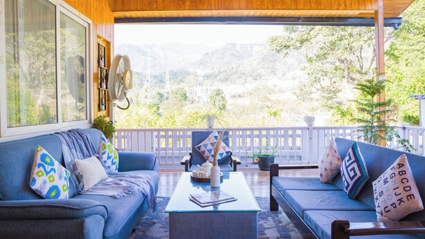 A covered porch with a blue sofa set, a glass coffee table, and a view of mountains. - Alaya Stays Baramasa 3 BHK Hill Retreat (Dehradun)