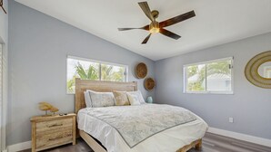 Un dormitorio con cama, mesita de noche, ventilador de techo y dos ventanas.