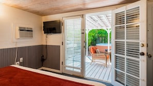 A room with a bed, a television mounted on the wall, an air conditioning unit, and a sliding glass door leading to an outdoor patio with a chair and a table. - Spacious Key West Conch Cottage with WiFi & Private Hot Tub - Dogs OK (Key West)