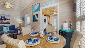 A cozy, well-lit room with a wooden ceiling, a dining table set with blue napkins, wicker chairs, and a view of the kitchen and laundry area. - Pool, Pet-friendly, Bicycles and 5 min walk to Beach (Key West)