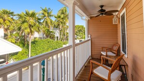 A wooden porch with a white railing, two wooden chairs with cushions, a ceiling fan, and a view of palm trees and a white house. - The Southernmost Comfort House — Private Pool + Waterfall | 5BR Truman Annex Estate | 28-Day Stay (Key West)