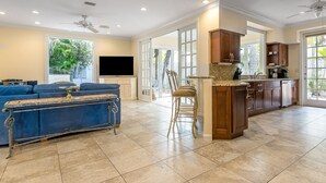 A spacious living area with a blue sofa, a wooden coffee table, and a kitchen with wooden cabinets and a tiled floor. - The Southernmost Comfort House — Private Pool + Waterfall | 5BR Truman Annex Estate | 28-Day Stay (Key West)