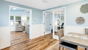 Una habitación con suelo de madera, una mesa de comedor con sillas, una pared azul claro y una puerta que da a otra estancia.