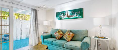 Ein Wohnzimmer mit einem türkisfarbenen Sofa, zwei gemusterten Kissen, einem gewebten Couchtisch und einem blauen Teppich mit Muster. An der Wand hängt ein Gemälde, auf einem Beistelltisch steht eine Lampe und man sieht durch das Fenster einen weißen Zaun und Grünflächen.