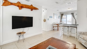 A living room with a large flat-screen TV, a wooden fish decoration, a dining area with a white table and chairs, and a kitchen with a bar counter. - Oceanfront Duplex with Beautiful Beach & Dock for Boat Access - Dogs OK (Long Key)